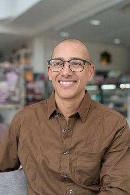 40 'lı yaşlarda kel bir adam kafeteryada oturuyor, günlük gömlek ve gözlük takıyor. Kendinden emin ve mutlu bir ifade. Kentsel yaşam tarzı, modern adam, rahat ortamda yetişkin portresi.