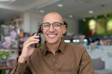 Gözlüklü kel Latin adam kafeteryada akıllı telefon kullanarak oturuyor. Sıradan bir gömlek giyiyor, gülümsüyor ve kendinden emin. Modern dijital yaşam tarzı, iletişim, mobil teknoloji, kentsel kahve dükkanı sahnesi.