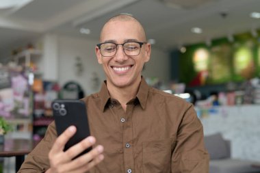 Gözlüklü kel Latin adam kafeteryada akıllı telefon kullanarak oturuyor. Sıradan bir gömlek giyiyor, gülümsüyor ve kendinden emin. Modern dijital yaşam tarzı, iletişim, mobil teknoloji, kentsel kahve dükkanı sahnesi.