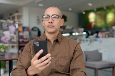 Gözlüklü kel Latin adam kafeteryada akıllı telefon kullanarak oturuyor. Sıradan bir gömlek giyiyor, gülümsüyor ve kendinden emin. Modern dijital yaşam tarzı, iletişim, mobil teknoloji, kentsel kahve dükkanı sahnesi.