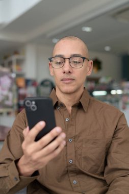 Gözlüklü kel Latin adam kafeteryada akıllı telefon kullanarak oturuyor. Sıradan bir gömlek giyiyor, gülümsüyor ve kendinden emin. Modern dijital yaşam tarzı, iletişim, mobil teknoloji, kentsel kahve dükkanı sahnesi.