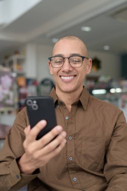 Gözlüklü kel Latin adam kafeteryada akıllı telefon kullanarak oturuyor. Sıradan bir gömlek giyiyor, gülümsüyor ve kendinden emin. Modern dijital yaşam tarzı, iletişim, mobil teknoloji, kentsel kahve dükkanı sahnesi.