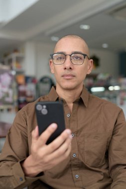 Gözlüklü kel Latin adam kafeteryada akıllı telefon kullanarak oturuyor. Sıradan bir gömlek giyiyor, gülümsüyor ve kendinden emin. Modern dijital yaşam tarzı, iletişim, mobil teknoloji, kentsel kahve dükkanı sahnesi.