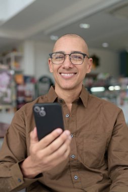 Gözlüklü kel Latin adam kafeteryada akıllı telefon kullanarak oturuyor. Sıradan bir gömlek giyiyor, gülümsüyor ve kendinden emin. Modern dijital yaşam tarzı, iletişim, mobil teknoloji, kentsel kahve dükkanı sahnesi.