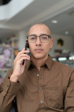 Gözlüklü kel Latin adam kafeteryada akıllı telefon kullanarak oturuyor. Sıradan bir gömlek giyiyor, gülümsüyor ve kendinden emin. Modern dijital yaşam tarzı, iletişim, mobil teknoloji, kentsel kahve dükkanı sahnesi.