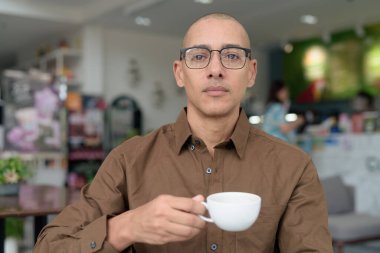 40 'lı yaşlarda kel bir adam kafeteryada oturuyor, günlük gömlek ve gözlük takıyor. Kendinden emin ve mutlu bir ifade. Kentsel yaşam tarzı, modern adam, rahat ortamda yetişkin portresi.