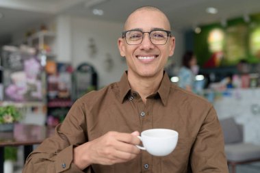 40 'lı yaşlarda kel bir adam kafeteryada oturuyor, günlük gömlek ve gözlük takıyor. Kendinden emin ve mutlu bir ifade. Kentsel yaşam tarzı, modern adam, rahat ortamda yetişkin portresi.