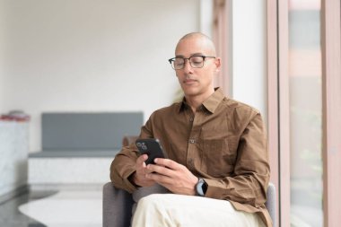 Gözlüklü kel Latin adam kafeteryada akıllı telefon kullanarak oturuyor. Sıradan bir gömlek giyiyor, gülümsüyor ve kendinden emin. Modern dijital yaşam tarzı, iletişim, mobil teknoloji, kentsel kahve dükkanı sahnesi.