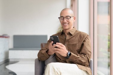 Gözlüklü kel Latin adam kafeteryada akıllı telefon kullanarak oturuyor. Sıradan bir gömlek giyiyor, gülümsüyor ve kendinden emin. Modern dijital yaşam tarzı, iletişim, mobil teknoloji, kentsel kahve dükkanı sahnesi.