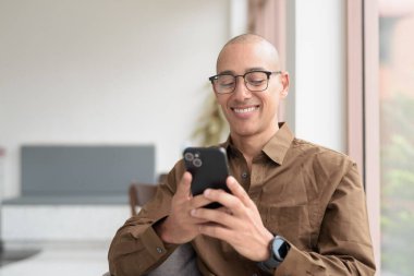 Gözlüklü kel Latin adam kafeteryada akıllı telefon kullanarak oturuyor. Sıradan bir gömlek giyiyor, gülümsüyor ve kendinden emin. Modern dijital yaşam tarzı, iletişim, mobil teknoloji, kentsel kahve dükkanı sahnesi.