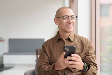 Gözlüklü kel Latin adam kafeteryada akıllı telefon kullanarak oturuyor. Sıradan bir gömlek giyiyor, gülümsüyor ve kendinden emin. Modern dijital yaşam tarzı, iletişim, mobil teknoloji, kentsel kahve dükkanı sahnesi.