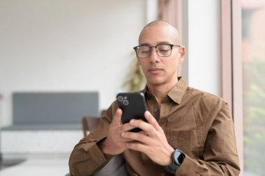 Gözlüklü kel Latin adam kafeteryada akıllı telefon kullanarak oturuyor. Sıradan bir gömlek giyiyor, gülümsüyor ve kendinden emin. Modern dijital yaşam tarzı, iletişim, mobil teknoloji, kentsel kahve dükkanı sahnesi.