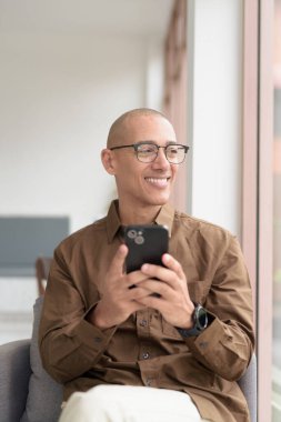 Gözlüklü kel Latin adam kafeteryada akıllı telefon kullanarak oturuyor. Sıradan bir gömlek giyiyor, gülümsüyor ve kendinden emin. Modern dijital yaşam tarzı, iletişim, mobil teknoloji, kentsel kahve dükkanı sahnesi.