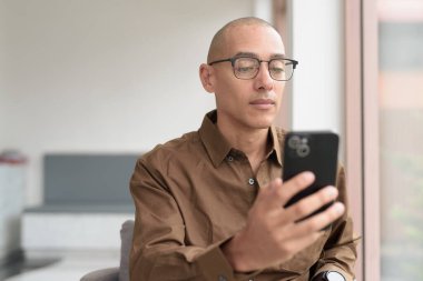 Gözlüklü kel Latin adam kafeteryada akıllı telefon kullanarak oturuyor. Sıradan bir gömlek giyiyor, gülümsüyor ve kendinden emin. Modern dijital yaşam tarzı, iletişim, mobil teknoloji, kentsel kahve dükkanı sahnesi.