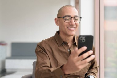 Gözlüklü kel Latin adam kafeteryada akıllı telefon kullanarak oturuyor. Sıradan bir gömlek giyiyor, gülümsüyor ve kendinden emin. Modern dijital yaşam tarzı, iletişim, mobil teknoloji, kentsel kahve dükkanı sahnesi.