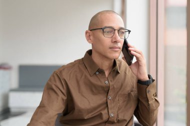 Gözlüklü kel Latin adam kafeteryada akıllı telefon kullanarak oturuyor. Sıradan bir gömlek giyiyor, gülümsüyor ve kendinden emin. Modern dijital yaşam tarzı, iletişim, mobil teknoloji, kentsel kahve dükkanı sahnesi.