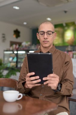 Rahat bir kafede otururken 40 'lı yaşlarındaki kel bir adamın tablet tabletini kullanması. Gözlük takıyor ve sıradan bir gömlek giyiyor. Teknoloji, dijital alışkanlıklar ve eğlence yaşam tarzı teması.