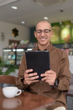 Rahat bir kafede otururken 40 'lı yaşlarındaki kel bir adamın tablet tabletini kullanması. Gözlük takıyor ve sıradan bir gömlek giyiyor. Teknoloji, dijital alışkanlıklar ve eğlence yaşam tarzı teması.