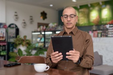 Rahat bir kafede otururken 40 'lı yaşlarındaki kel bir adamın tablet tabletini kullanması. Gözlük takıyor ve sıradan bir gömlek giyiyor. Teknoloji, dijital alışkanlıklar ve eğlence yaşam tarzı teması.