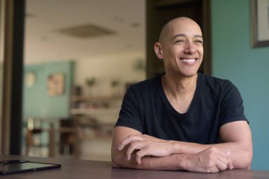 Kütüphanede oturmuş pencereden bakarken gülümseyen, günlük siyah tişörtlü mutlu, kel bir İspanyol. Kitap rafları ve rahat atmosferi olan parlak modern kapalı alan.