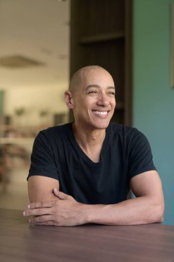 Kütüphanede oturmuş pencereden bakarken gülümseyen, günlük siyah tişörtlü mutlu, kel bir İspanyol. Kitap rafları ve rahat atmosferi olan parlak modern kapalı alan.