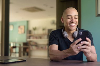 Kütüphanede pencerenin önünde oturan, günlük siyah tişörtlü kel adam akıllı telefon kullanıyor. Kitap raflarıyla, akademik atmosferiyle ve rahat ruh haliyle modern iç mekanlar..