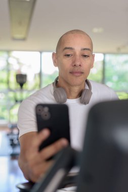 Modern spor salonunun içinde telefon kullanırken sabit egzersiz bisikleti üzerinde çalışan kel bir İspanyol. Güç, kardiyo ve motivasyonu gösteren odaklanmış fitness ve egzersiz konsepti.