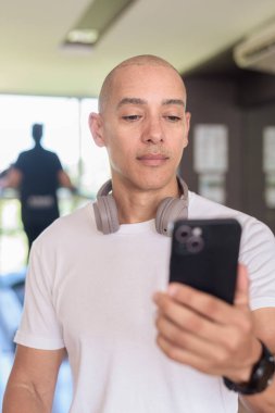 Spor salonundaki kel Latin adam setler arasında telefon kullanıyor. Dijital fitness izleme, sağlık uygulamaları, kişisel eğitim alışkanlıkları ve modern egzersiz yaşam tarzını temsil etmek.