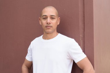 Beyaz tişörtlü, kel Latin bir adam duvara yaslanmış. Minimalist, stil özgüven, kimlik ve sağlık yaşam tarzını vurguluyor.