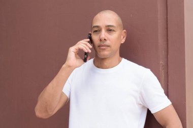 Beyaz tişörtlü, kel Latin bir adam duvara yaslanmış. Minimalist, stil özgüven, kimlik ve sağlık yaşam tarzını vurguluyor.