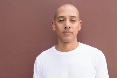 Beyaz tişörtlü, kel Latin bir adam duvara yaslanmış. Minimalist, stil özgüven, kimlik ve sağlık yaşam tarzını vurguluyor.