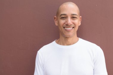 Beyaz tişörtlü, kel Latin bir adam duvara yaslanmış. Minimalist, stil özgüven, kimlik ve sağlık yaşam tarzını vurguluyor.