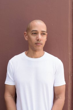 Beyaz tişörtlü, kel Latin bir adam duvara yaslanmış. Minimalist, stil özgüven, kimlik ve sağlık yaşam tarzını vurguluyor.