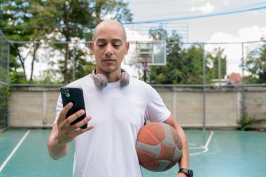 Spor giyim bölümündeki kel adam basketbol sahasında akıllı telefon kullanıyor. Kentsel fitness ve sağlıklı yaşam tarzı konsepti modern teknoloji ile atletik eğitimi harmanlıyor.