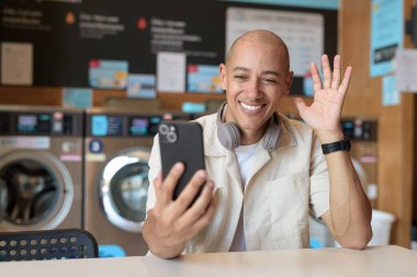 Günlük kıyafetli kel bir İspanyol, çamaşırhanede akıllı telefon kullanarak oturuyor. Arka planda çamaşır makineleri olan günlük şehir hayatı.