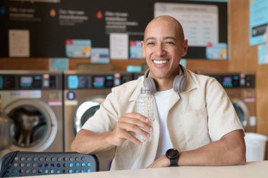 Günlük kıyafetli kel bir adam çamaşırhanede oturmuş su şişesinden içiyor. Arka planda çamaşır makineleri olan şehir günlük yaşam tarzı.