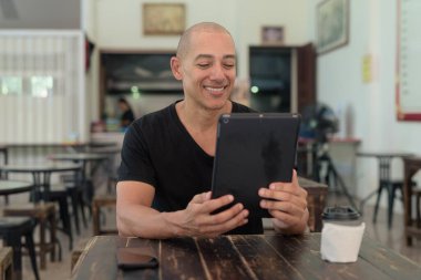 Siyah tişört giyen kel Latin bir adamın portresi, basit bir açık kahve dükkanında tek başına oturuyor.