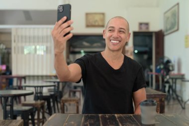 Siyah tişört giyen kel Latin bir adamın portresi, basit bir açık kahve dükkanında tek başına oturuyor.