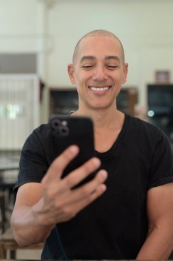 Siyah tişört giyen kel Latin bir adamın portresi, basit bir açık kahve dükkanında tek başına oturuyor.