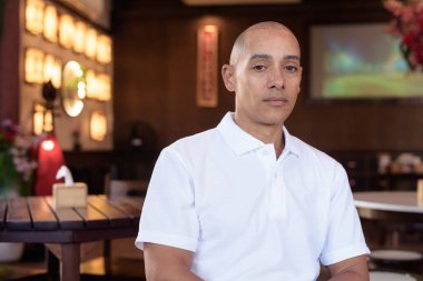 Beyaz polo tişörtlü kel Latin bir adamın portresi, şık bir Çin tarzı kahve dükkanında oturuyor, ev ortamında kendine güveni tam görünüyor..