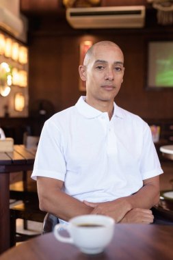 Beyaz polo tişörtlü kel Latin bir adamın portresi, şık bir Çin tarzı kahve dükkanında oturuyor, ev ortamında kendine güveni tam görünüyor..