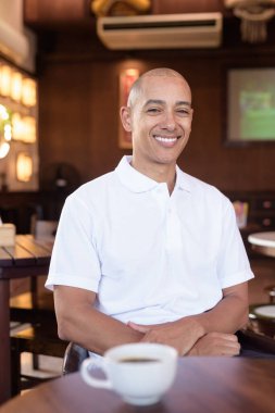 Beyaz polo tişörtlü kel Latin bir adamın portresi, şık bir Çin tarzı kahve dükkanında oturuyor, ev ortamında kendine güveni tam görünüyor..