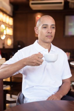 Beyaz polo tişörtlü kel Latin bir adamın portresi, şık bir Çin tarzı kahve dükkanında oturuyor, ev ortamında kendine güveni tam görünüyor..