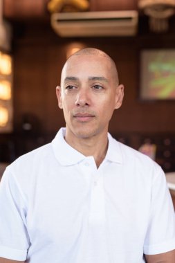 Beyaz polo tişörtlü kel Latin bir adamın portresi, şık bir Çin tarzı kahve dükkanında oturuyor, ev ortamında kendine güveni tam görünüyor..