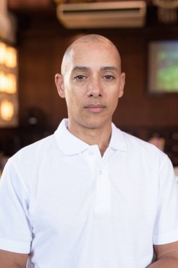 Beyaz polo tişörtlü kel Latin bir adamın portresi, şık bir Çin tarzı kahve dükkanında oturuyor, ev ortamında kendine güveni tam görünüyor..