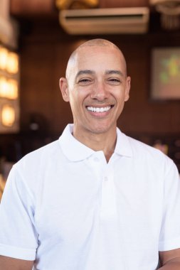 Beyaz polo tişörtlü kel Latin bir adamın portresi, şık bir Çin tarzı kahve dükkanında oturuyor, ev ortamında kendine güveni tam görünüyor..