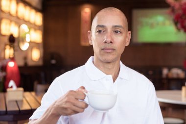 Beyaz polo tişörtlü kel Latin bir adamın portresi, şık bir Çin tarzı kahve dükkanında oturuyor, ev ortamında kendine güveni tam görünüyor..