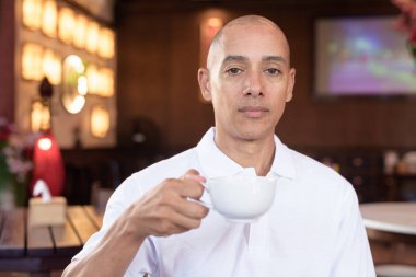 Beyaz polo tişörtlü kel Latin bir adamın portresi, şık bir Çin tarzı kahve dükkanında oturuyor, ev ortamında kendine güveni tam görünüyor..