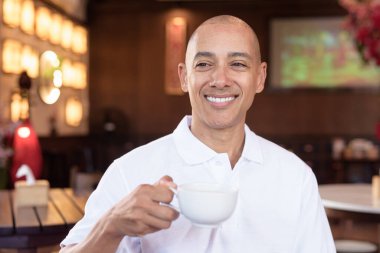 Beyaz polo tişörtlü kel Latin bir adamın portresi, şık bir Çin tarzı kahve dükkanında oturuyor, ev ortamında kendine güveni tam görünüyor..