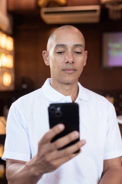 Beyaz polo tişörtlü kel Latin bir adamın portresi, şık bir Çin tarzı kahve dükkanında oturuyor, ev ortamında kendine güveni tam görünüyor..